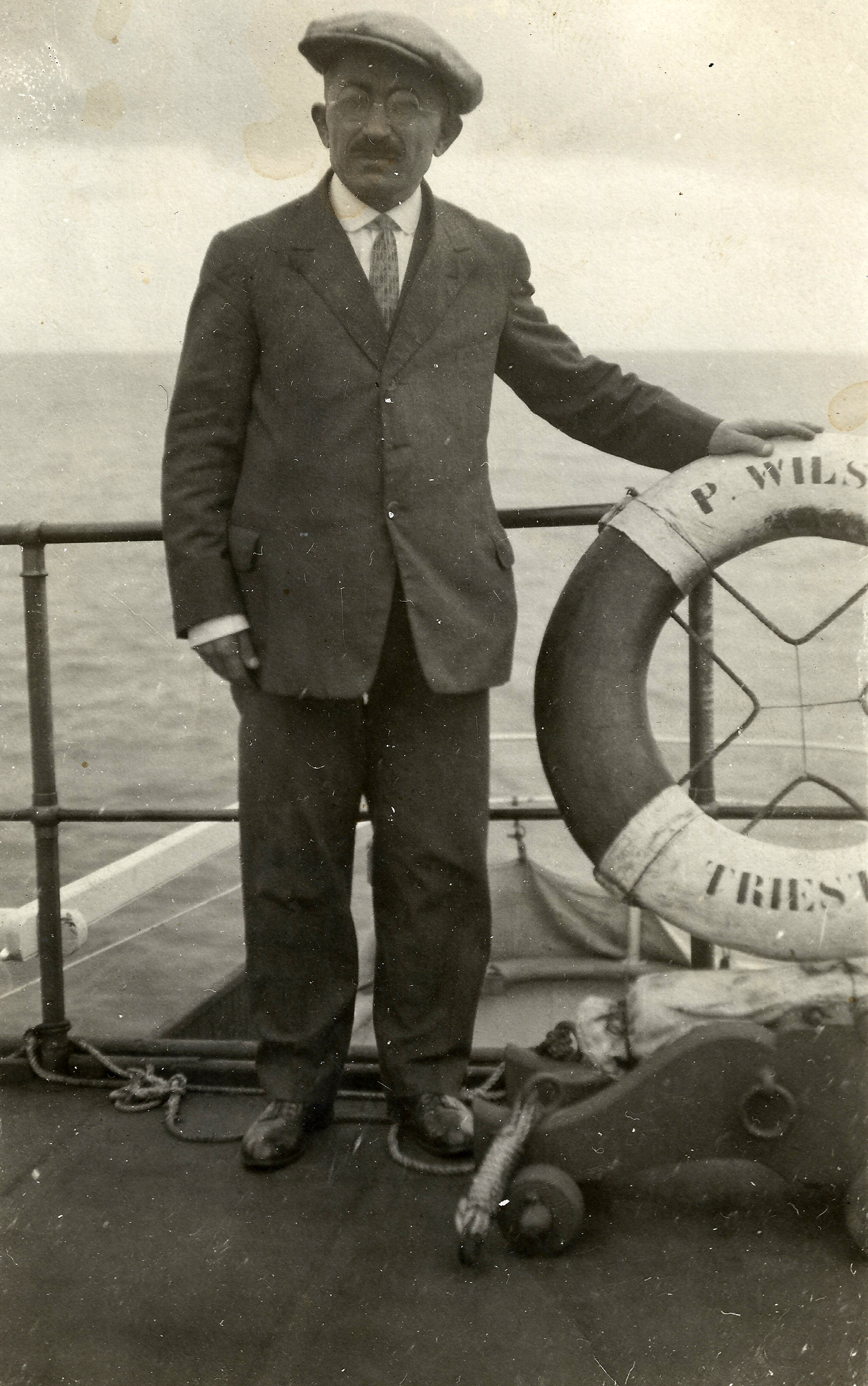 homem com vestes formais dentro de um barco, ao seu lado uma boia de duas cores, escura e clara. Foto preto e branca, ao fundo pode se visualizar o oceano