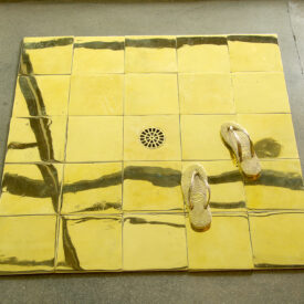 azulejos amarelos um ao lado do outro, ao centro um ralo, ao lado direito inferior dois pés de sandálias