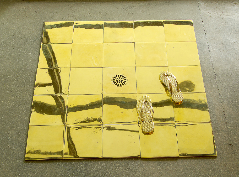azulejos amarelos um ao lado do outro, ao centro um ralo, ao lado direito inferior dois pés de sandálias