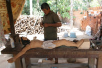 santeiro esculpindo jesus na cruz, homem em segundo plano ao lado da mesa ao fundo possuimos materia prima dos santeiros