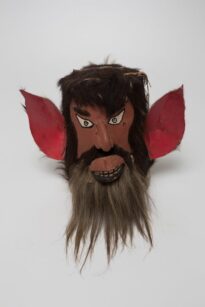 máscara, antropomorfo, orelhas de doende, barba de humano, sorrico com dentes aparentes, pele parda, olhos triangulares, cabelo marrom com core masculino, bigode junto com costeleta e barba