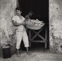 Homem adulto por volta dos 40 anos em pé a um passo do portal de entrada do edifício. Ele tem porte médio, possui cabelos escuros e bigode grosso, usa roupas claras e está com os pés descalços. Este homem segura com as duas mãos ao seu lado esquerdo um grande cesto de pães
