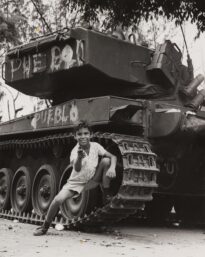 Garoto apoiado em tanque de guerra, garoto faz sinal de arma apontando o indicador para o fotografo, ele está com a perna esquerda dobrada encostada no pneu do tanque, ele expressa um sorriso de mostrar os dentes. No tanque temos pixadas as palavras "PUEBLO" na cor branca, a escrita está presente na parte superior e inferior do veículo