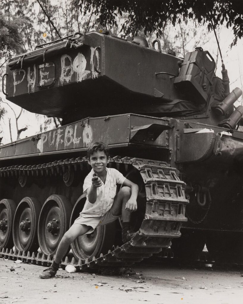 Garoto apoiado em tanque de guerra, garoto faz sinal de arma apontando o indicador para o fotografo, ele está com a perna esquerda dobrada encostada no pneu do tanque, ele expressa um sorriso de mostrar os dentes. No tanque temos pixadas as palavras "PUEBLO" na cor branca, a escrita está presente na parte superior e inferior do veículo