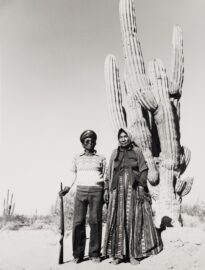 casal de mexicanos em primeiro plano, em segundo plano exemplar cactáceas, em terceiro plano o deserto e o céu. A mulher usa um vestido e um turbante tradicional. O homem usa calças escuras blusa clara e chapéu quepe, em sua mão direita ele segura uma espingarda pelo cano, a outra extremidade está apoiada ao solo.