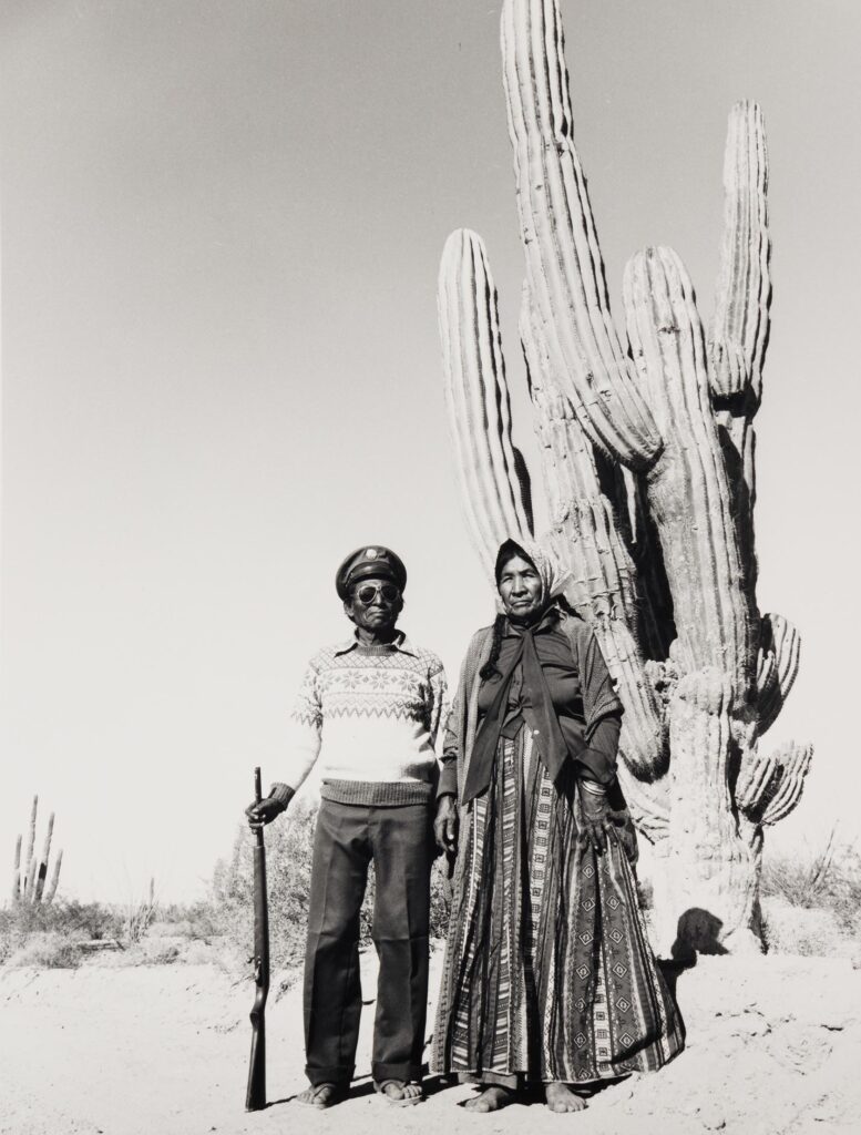 casal de mexicanos em primeiro plano, em segundo plano exemplar cactáceas, em terceiro plano o deserto e o céu. A mulher usa um vestido e um turbante tradicional. O homem usa calças escuras blusa clara e chapéu quepe, em sua mão direita ele segura uma espingarda pelo cano, a outra extremidade está apoiada ao solo.