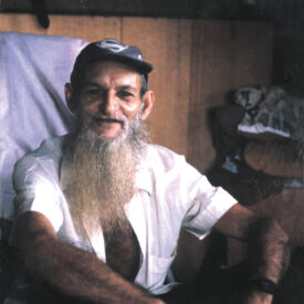 Padrinho Sebastião sentado no chão, sorrindo de chapéu e camiseta branca de meia manga.Ao fundo parede de madeira.