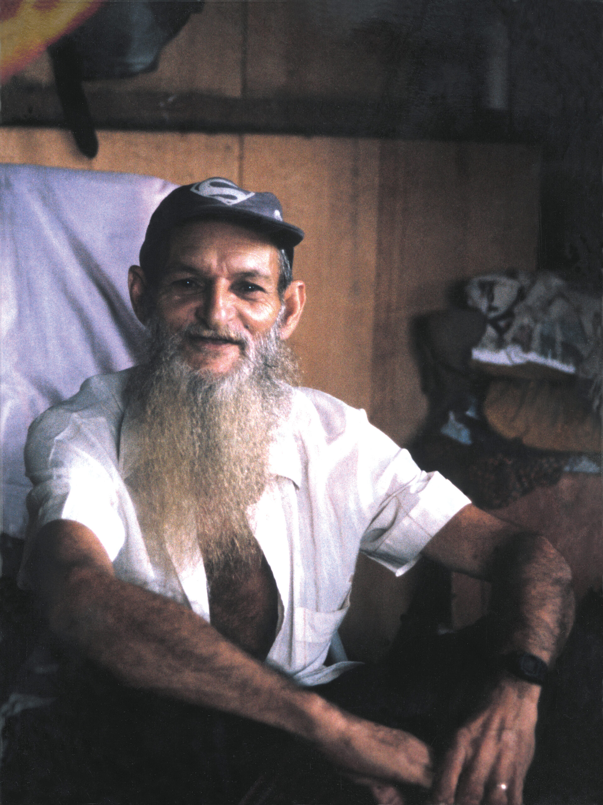 Padrinho Sebastião sentado no chão, sorrindo de chapéu e camiseta branca de meia manga.Ao fundo parede de madeira.