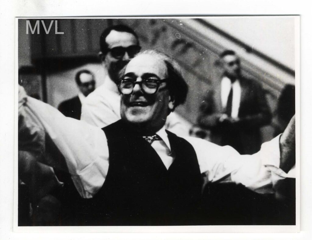 Em primeiro plano Villa Lobos, ele está de braços erguidos, usa um óculos e tem aberto grande sorriso.
