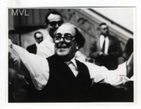 Em primeiro plano Villa Lobos, ele está de braços erguidos, usa um óculos e tem aberto grande sorriso.