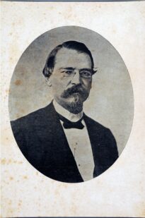 Dentro de oval, busto de figura masculina levemente voltado à direita, cabelos partidos na altura da orelha, usa pince-nez, bigode e cavanhaque. Trajando indumentária escura com gola batida, camisa branca com gola de padre e gravata borboleta escura. Fundo cinza.